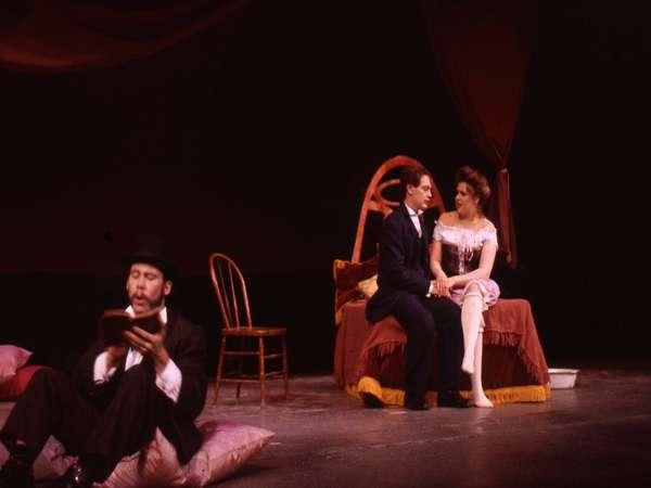 A theater stage scene with a man and woman seated on a bed, the man in a dark suit and the woman in a corset. A second man sits on the floor reading, wearing a hat and dark clothing. The background is dimly lit, suggesting an intimate setting.