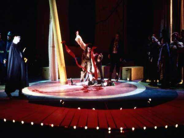 A theatrical performance on stage features a dramatic scene with a person kneeling in the center, raising red ribbons. The stage is circular, surrounded by spotlights, with other performers standing around the edges in dim lighting.