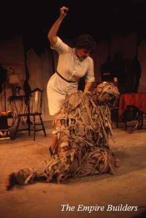 A woman stands on a stage holding a raised object, poised to strike. In front of her, a person is covered in tattered fabric, kneeling. The setting suggests a theatrical performance of "The Empire Builders.