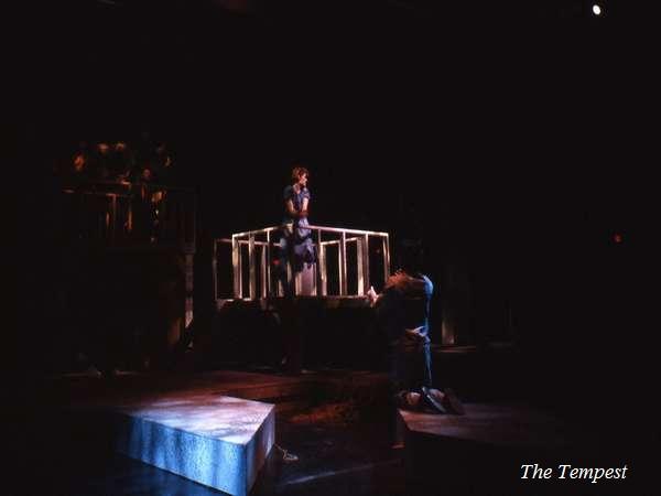A dimly lit stage scene from a theater production of "The Tempest." Two actors perform, one standing on a structure resembling a platform or cage, and another kneeling. The stage lighting is dramatic and shadowed.