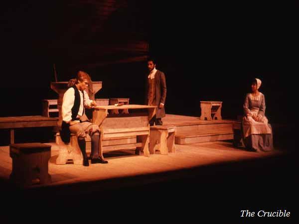 A dimly lit stage scene from "The Crucible" features three actors in period costumes. One sits thoughtfully at a wooden table, another stands nearby, and a third sits on a stool in a bonnet and apron. The setting is minimalistic with wooden props.