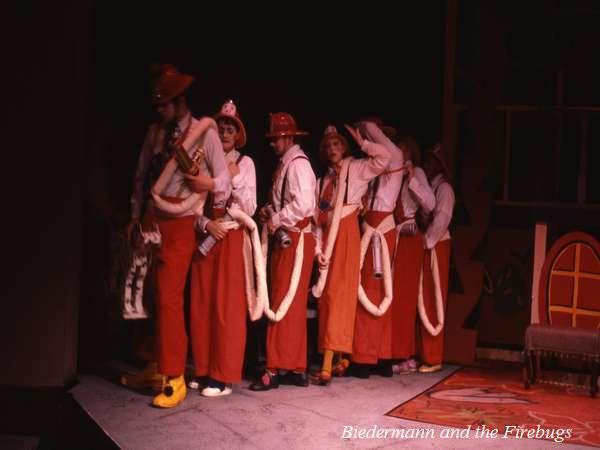 A group of actors dressed as firefighters stand in a line on stage. They wear red pants, white shirts, and red helmets, with white ropes slung over their shoulders. The stage is minimally lit, with a partial view of a set to the right.