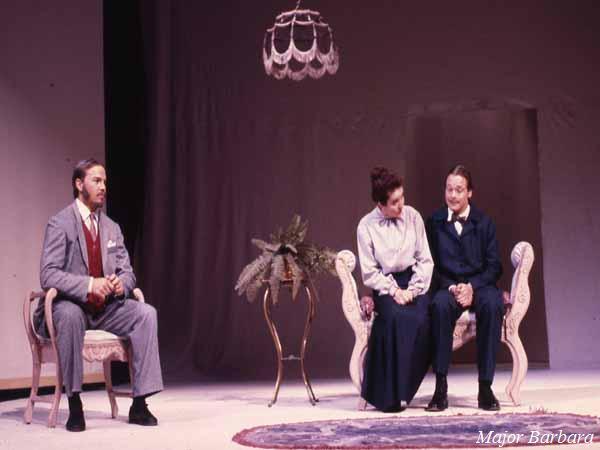 Three actors perform a scene on stage in a play titled "Major Barbara." Two women sit on a loveseat, while a man sits opposite them. A plant stands on a small table center stage. They are under a chandelier in a simple room setting.