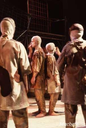 A group of actors in tattered, earth-toned costumes, with their heads covered, stand on stage. One actor has their hand raised to their face. The stage is dimly lit, and a cage-like structure is visible in the background. The word "Macbeth" is in the corner.