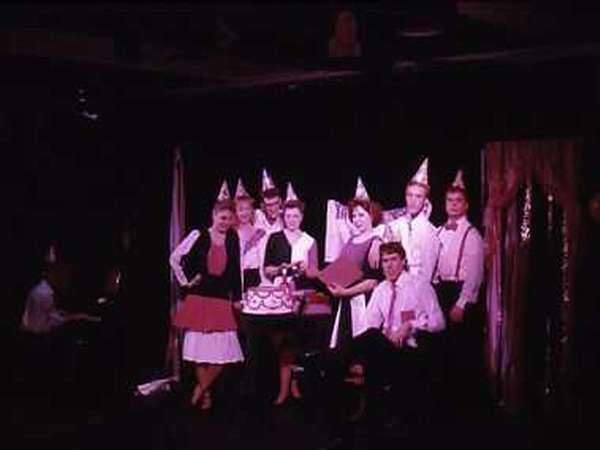 A group of eight people wearing party hats stand on a dimly lit stage. Some are holding what appears to be a large, decorated cake. They are dressed in formal or semi-formal attire, with some wearing vests and skirts.