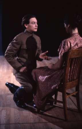 A person in a military uniform kneels on the wooden floor, gesturing while talking to another seated person in a patterned dress. The background is dark, emphasizing the interaction between them.