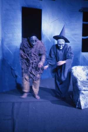 Two people in a dimly lit room wearing theatrical costumes. One wears tattered robes and a mask, while the other wears a pointed hat and a mask. Both stand near a piece of furniture with a backdrop resembling a wall with windows.