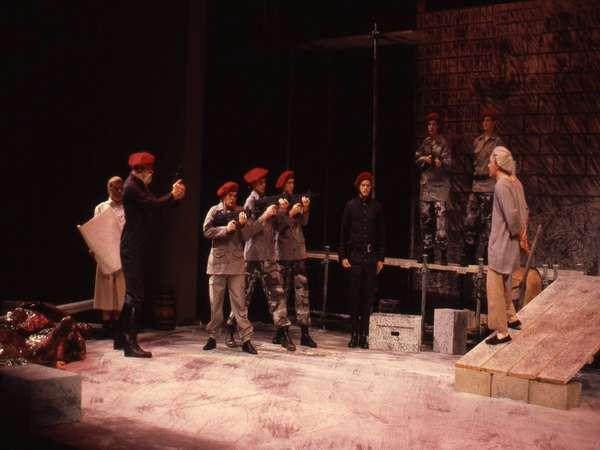 Stage performance depicting a dramatic scene with actors in military attire pointing guns. A figure stands on a platform facing them, while others observe. The setting includes dark, textured walls and various props, suggesting a tense narrative.