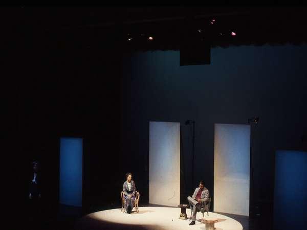 Two people sit on a dimly lit stage with minimal props. One is on the left in a dark outfit, and the other is on the right in a light outfit. The background is dark with three illuminated panels. The setting suggests a theatrical performance or play.