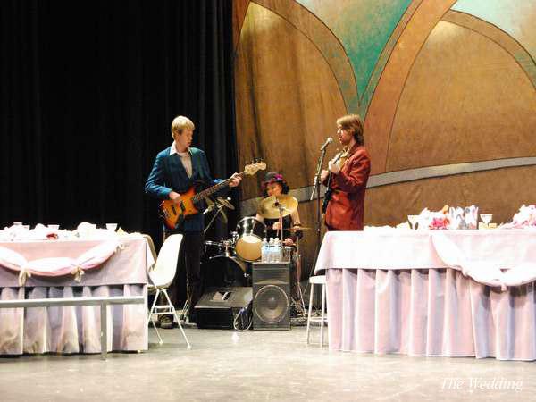 Three musicians perform on a stage. One plays an electric guitar, another is on drums, and the third holds a microphone. The setting includes tables with pink tablecloths and decor, suggesting a formal event.