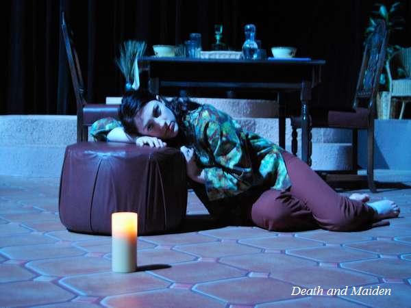 A person in a patterned shirt and maroon pants rests their head on a brown cushion, seated on a tiled floor. A lit candle is in the foreground. Behind is a table set with glasses and plates. The text "Death and Maiden" is in the bottom right corner.
