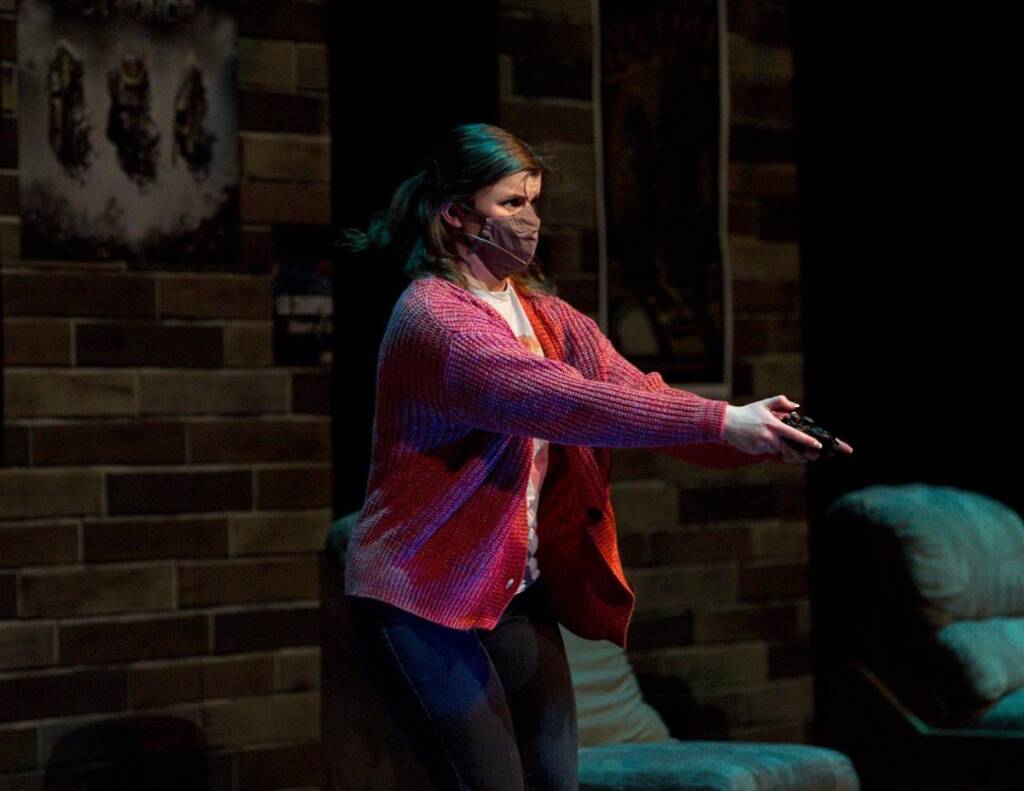 A woman wearing a red cardigan and a face mask holds a controller and stands in a dimly lit room with brick walls and posters. She appears focused, with a couch visible in the background.
