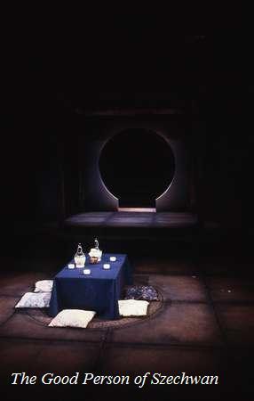 A dimly lit stage with a circular opening in the background. A blue table with glass bottles and several cushions is in the center. The play's title, "The Good Person of Szechwan," is displayed at the bottom.