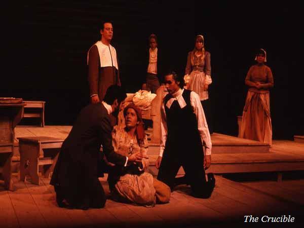 Theater performance of "The Crucible" showing actors on a dimly lit wooden stage. One actor kneels in front of a seated woman, while others stand in Puritan costumes. A sense of tension and drama is evident in their poses and expressions.