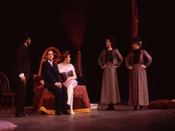 Five actors on stage perform a scene set in historical costumes. Two men in suits, one seated on a bed with a woman in a white dress, and two women in long dresses stand nearby, creating a dramatic atmosphere.