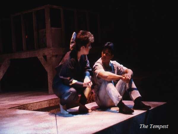 Two actors perform a scene on stage from "The Tempest." One kneels, engaged in conversation, while the other sits cross-legged. The set features simple wooden structures, and the lighting creates a dramatic ambiance.