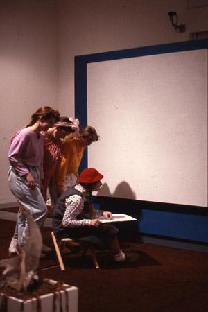 A group of four children engage with a large white screen. One child in a red hat sits at a small table, drawing, while the others, dressed in colorful clothes, stand nearby, watching attentively. A potted plant is visible in the foreground.