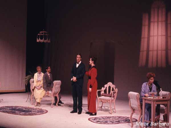 Five people are on a stage set with simplistic furniture. A man and woman stand in the center, while others are seated around them. The scene is set to resemble a living room. Text reading "Major Barbara" is in the lower right corner.