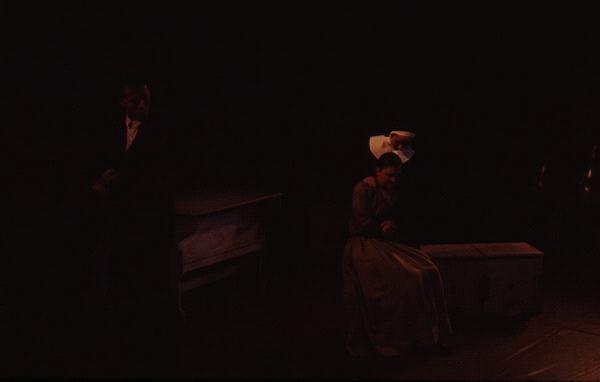 A dimly lit stage features a man standing on the left and a woman sitting on a trunk on the right. She appears to be deep in thought or sadness. A white chair is visible behind her. The overall mood is somber and dramatic.