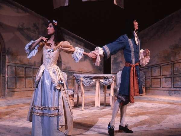 Two actors in period costumes perform on stage. The woman wears a long, ornate gown with a flower in her hair, while the man wears a blue coat with ruffled sleeves. They hold hands in a dramatic pose, with a stylized interior set as the backdrop.