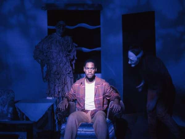 A person sits in a chair in a dimly lit room, lit with blue light, wearing casual clothes. Figures in masks and costumes stand behind and beside them, creating an eerie atmosphere.