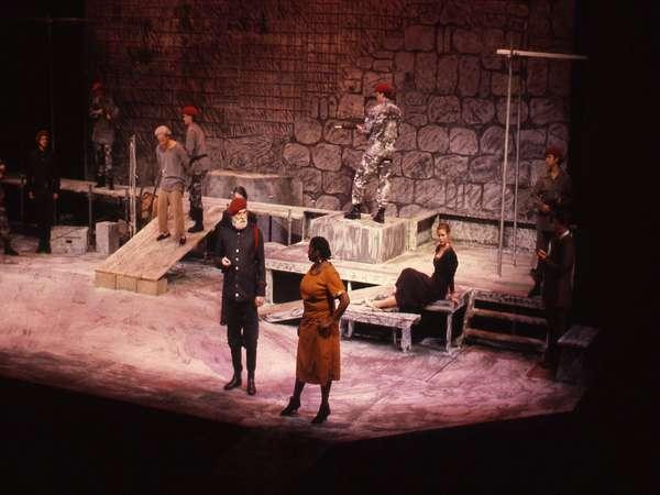 A theater stage with actors in costumes. The set has a stone wall backdrop, wooden platforms, and stairs. One actor in a long coat and red hat stands with another in a brown dress. Other actors are positioned around the stage, some seated.
