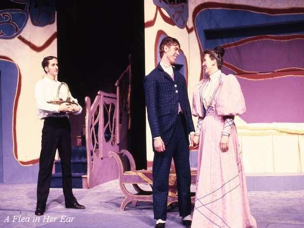 Three actors perform on stage in a play. Two, a man in a suit and a woman in a long purple dress, smile at each other. The third actor, dressed in white, stands aside holding a basket. The stage is decorated with colorful, abstract designs.