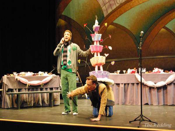 Two actors on stage in colorful costumes; one stands with a phone, wearing green pants and holding a cake prop. The other crouches beneath the cake, in front of a decorated table with a pastel backdrop and microphone.
