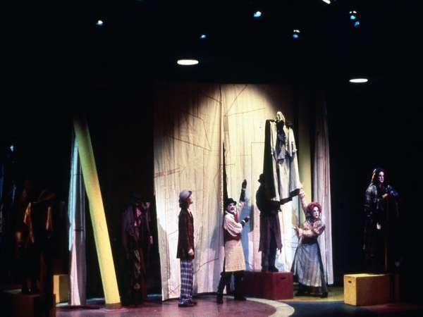 Stage performance with actors in colorful costumes standing by a curtain backdrop. One actor holds a large puppet or costume piece. The stage is dimly lit, with spotlights highlighting the performers. Brightly colored blocks are on the stage.