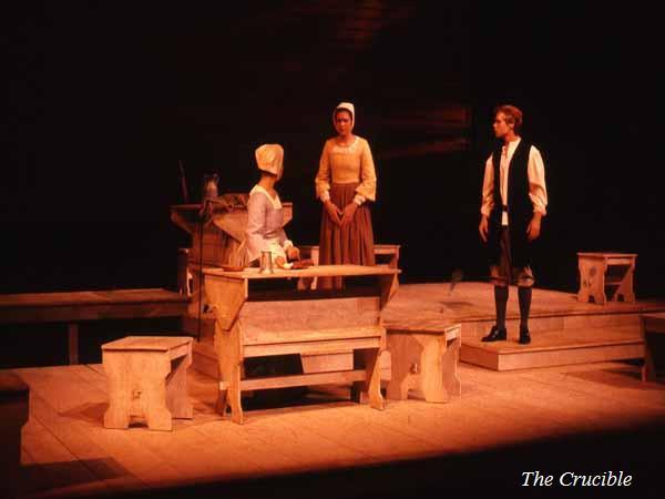 Two actors in period costumes on a stage set resembling a rustic room, with a wooden table and benches. The scene from "The Crucible" captures an intense moment, with one actor sitting and another standing, engaged in conversation.