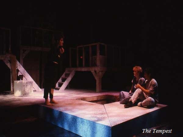 A dimly lit stage scene from "The Tempest" shows three actors. One stands on a platform, wearing dark clothing, while two others sit on a lower level, engaged in conversation. The set includes elevated platforms and a central opening.