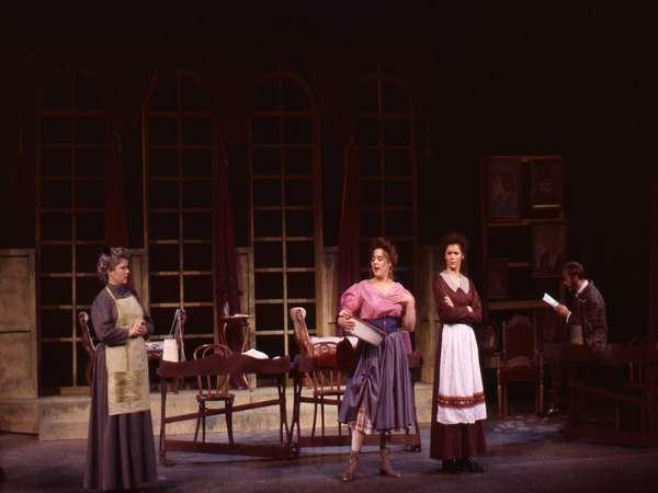 A theatrical scene with four actors in period costumes on stage. A woman in a pink blouse is speaking to another woman with arms crossed. Two other actors, a seated man and an older woman, look on. The set resembles a vintage dining room with large windows.