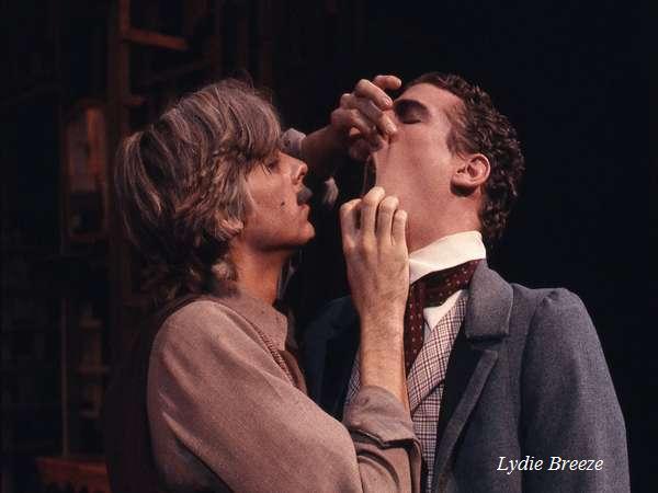 Two actors perform a dramatic scene on stage. One actor, in a brown shirt, holds the jaw of another actor, in a gray suit with a checkered vest, with his mouth open as if inspecting or treating him. The text "Lydie Breeze" is on the image.