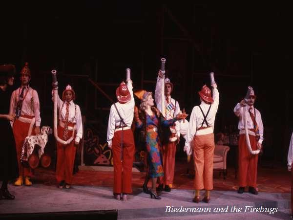 A stage performance of "Biedermann and the Firebugs" featuring actors in red pants, white shirts, and fire helmets, some holding hoses upward. A woman in a colorful dress stands among them, gesturing animatedly.