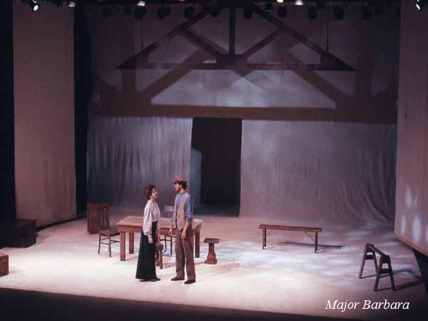 A minimalist stage set for "Major Barbara" with two actors in period costumes standing in the center. The backdrop features a simple wooden beam and doorframe, with scattered furniture around the stage.