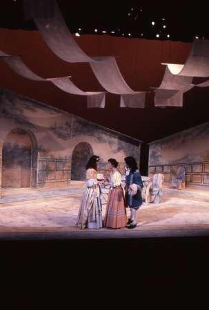 Three actors in period costumes perform on a dimly lit stage with a minimalistic set featuring painted walls and draped fabric overhead. The scene creates an atmosphere reminiscent of a historical drama.