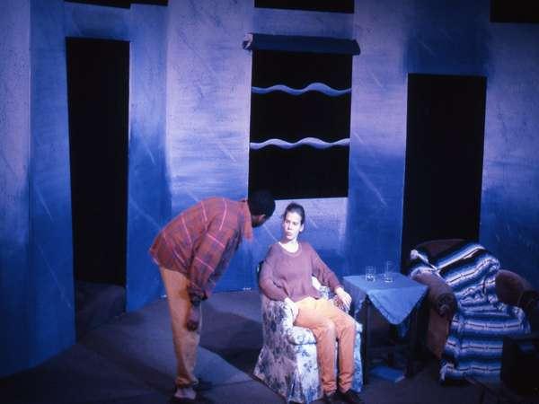 A person stands and leans toward another person sitting in a floral armchair on a stage set with a blue background. They appear to be engaged in a conversation. There is a round table covered with a striped cloth nearby.