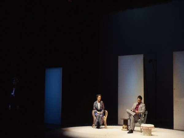 Two actors on stage are seated, surrounded by minimalistic set elements. The scene is dimly lit, with the spotlight on both individuals. One actor is dressed in a suit, while the other wears a patterned outfit.