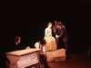 A dimly lit stage shows a person sitting at a table with a typewriter, another person sitting next to them. Two people are standing close, engaged in conversation. The seated person is wearing a long dress.