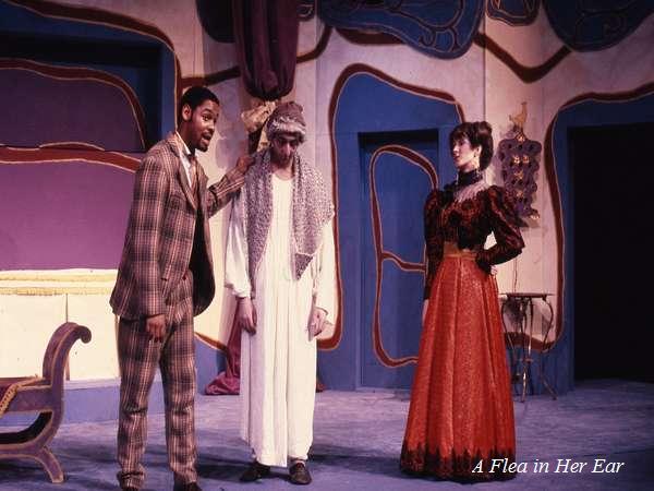 Three actors on stage perform a scene from a play titled "A Flea in Her Ear." One actor in a plaid suit talks animatedly, another in a robe and headscarf stands in the center, and a woman in a red Victorian dress looks on.