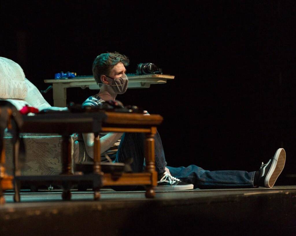 A person wearing a face mask sits on the floor of a dimly lit stage. They lean against a couch with scattered items, including a pair of headphones on a nearby table. The person is casually dressed in jeans and sneakers.