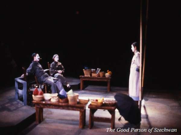 A theatrical scene featuring three actors on stage in dim lighting. Two actors sit on a bench with baskets around them, while a third stands nearby. The set is minimal, suggesting a simple setting. The text "The Good Person of Szechwan" is visible.