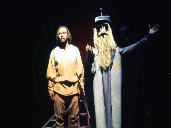 A person in worn clothing stands next to a tall figure with a long beard and a hat, dressed in a robe-like outfit, both on a dark stage. The scene is dimly lit, creating a dramatic atmosphere.