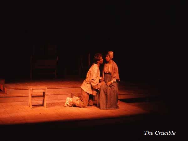 Two actors kneel on stage, illuminated by warm lighting, in a scene from "The Crucible." They are dressed in period clothing, with a simple wooden set and minimal props in the background.