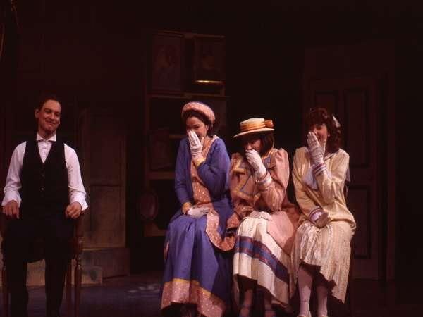 A man in a vest and tie sits on a chair, smiling. Next to him, three women in vintage dresses and hats cover their mouths with gloved hands, appearing amused. The setting resembles a theatrical stage with dim lighting and framed pictures on the wall.