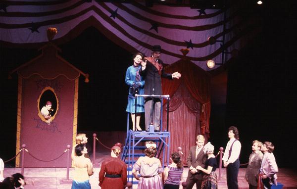 A stage performance with actors in 1930s-style costumes. A man and woman stand elevated, speaking to an audience. A painted backdrop featuring stripes and stars suggests a circus or carnival theme. Spectators watch from below.