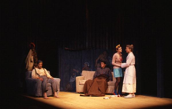 Four actors are on stage in a dimly lit theater scene. One is seated on the left in a chair. Two are in the center, one sitting on a sofa and another standing beside. The fourth stands on the right. The set includes worn furniture and a visible coat rack.