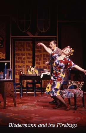 Two actors on stage in a dramatic scene from a play titled "Biedermann and the Firebugs." The woman is in a colorful dress, gesturing animatedly, while the man stands behind her with a surprised expression. The set is styled with vintage decor.