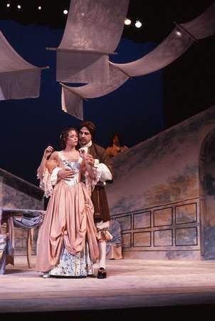 A man and woman in period costumes perform on stage. The woman wears a flowing dress and the man a rustic outfit. They are under draped fabric, suggesting a historical or romantic setting. The background features a painted scene.