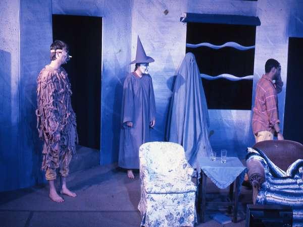 Four people in a dimly lit room performing on stage. One wears a shaggy costume, another a pointed hat and robe, the third is covered with a sheet, and the fourth is in casual clothes. Furniture includes an armchair and a small table with glasses.